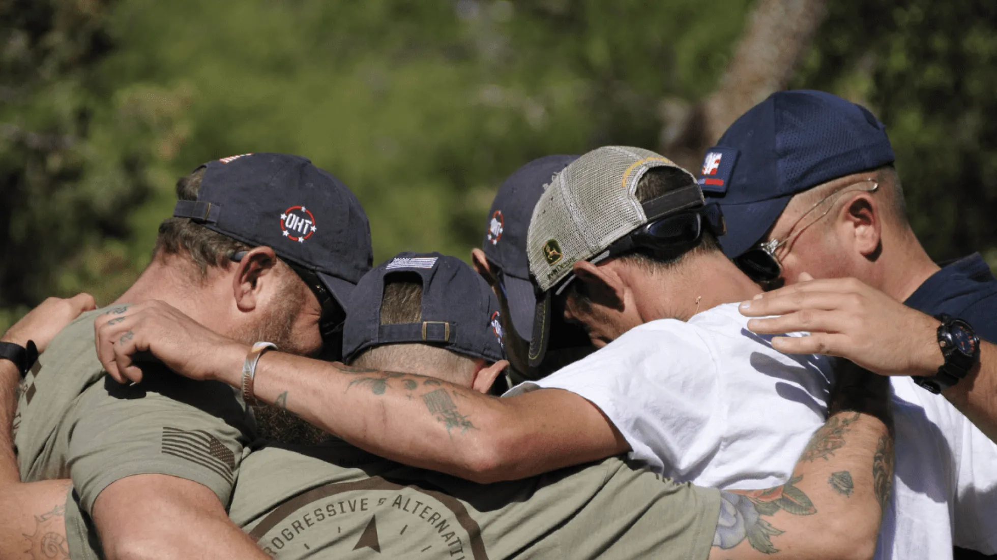 A group of men stand shoulder to shoulder and lock arms in a large group hug. Their faces are obscured.