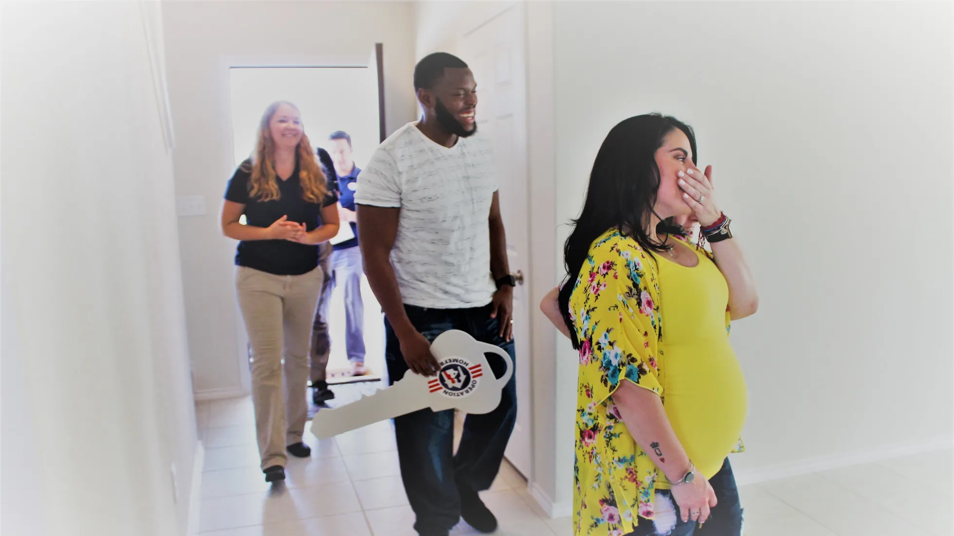 A mother-to-be gasps with joy as she enters her new home. Her husband follows her, holding a large key adorned with the Operation Homefront logo.
