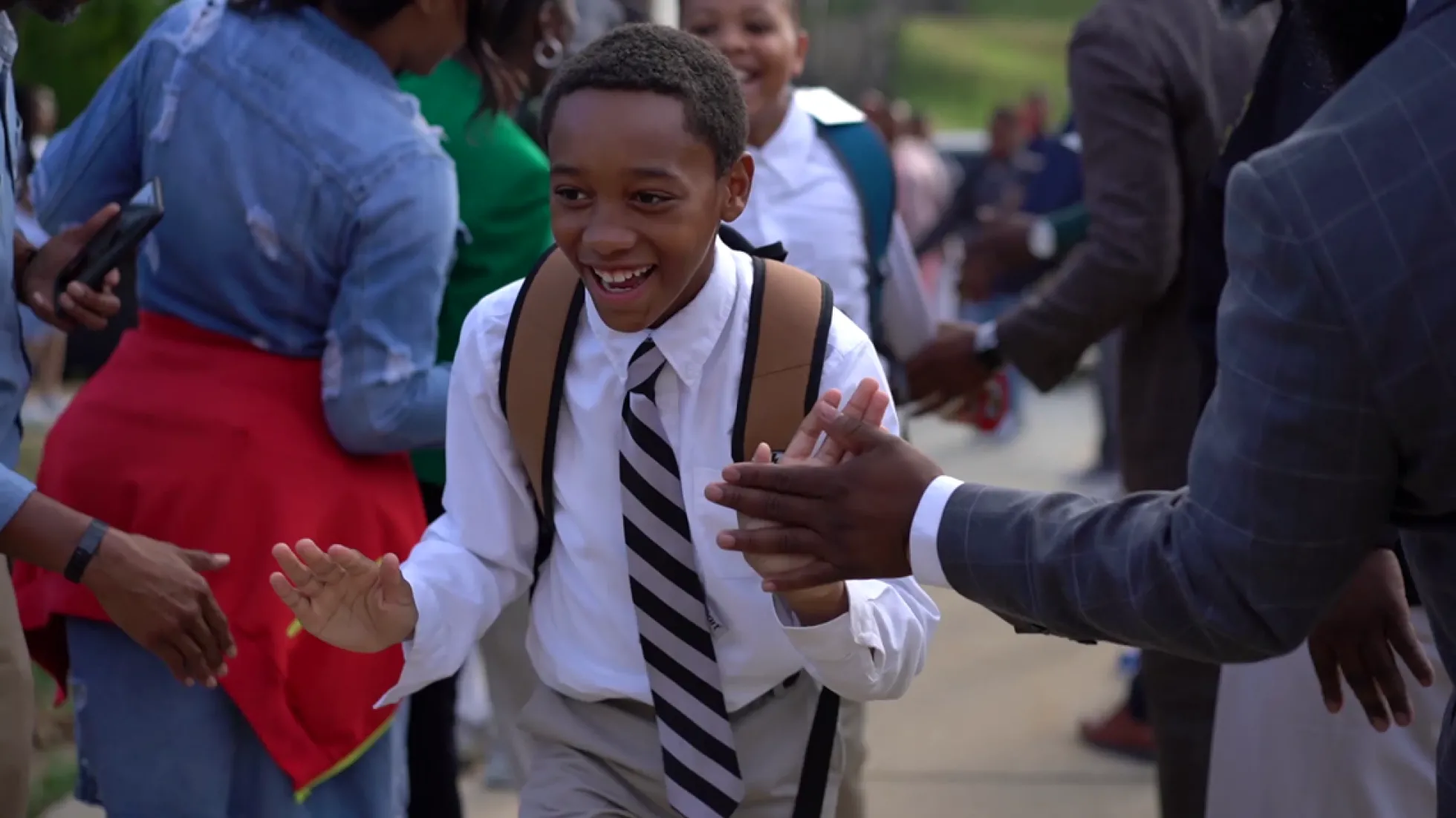 An elementary school student walks through a tunnel of adults who are cheering him on. The students and adults are wearing business attire.