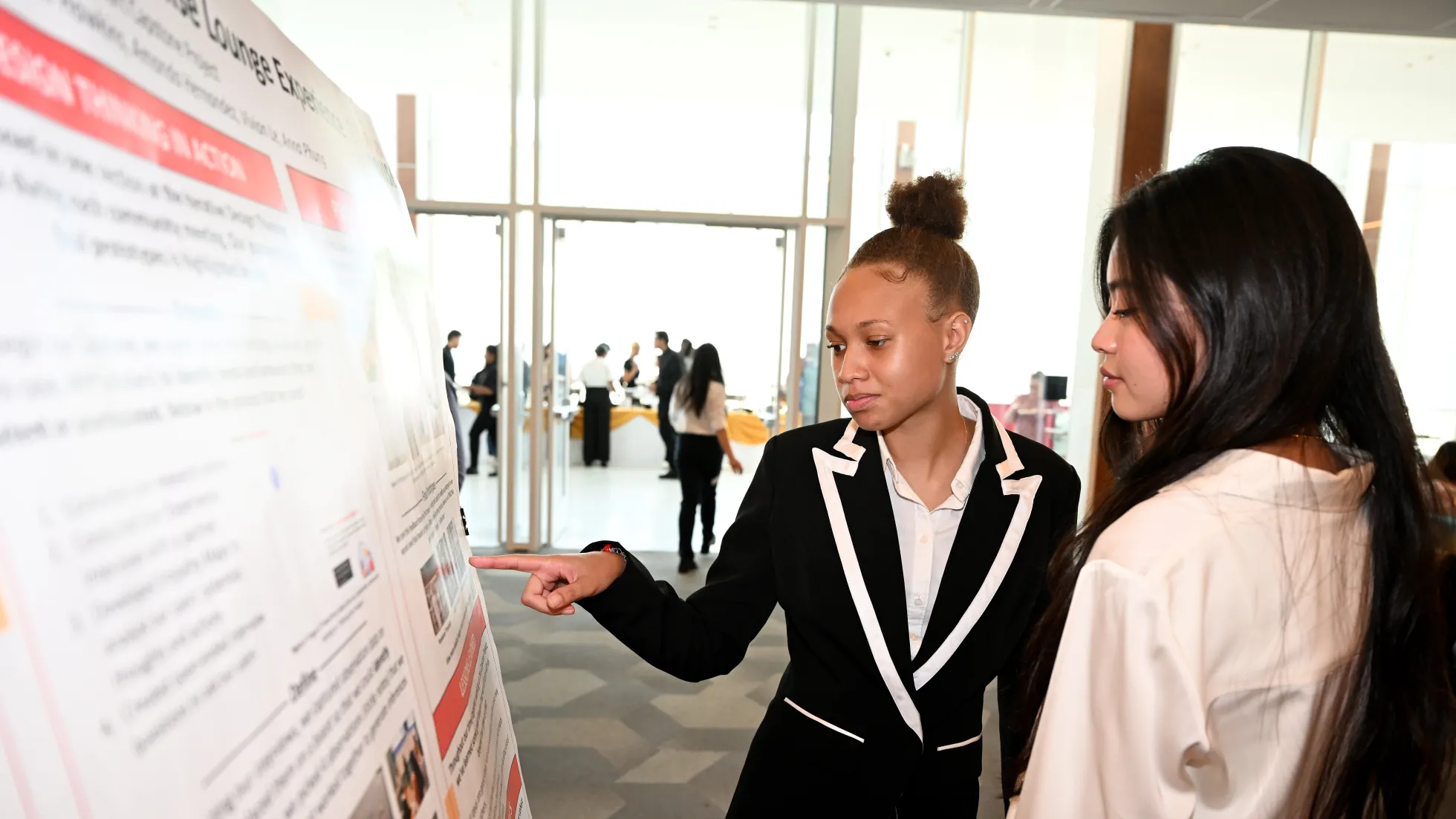 A student in business attire explains a presentation to another student while pointing at a poster board.