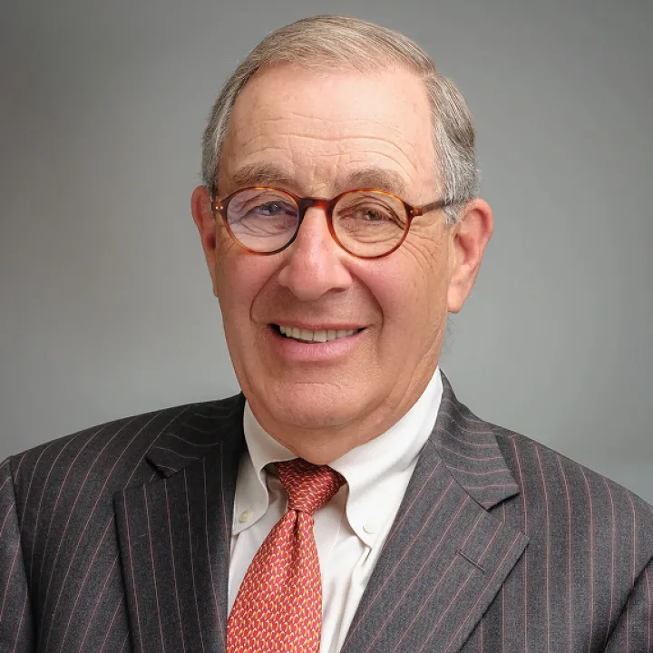 A professional headshot of Larry Nussdorf. He wears glasses, a grey blazer and an orange tie.