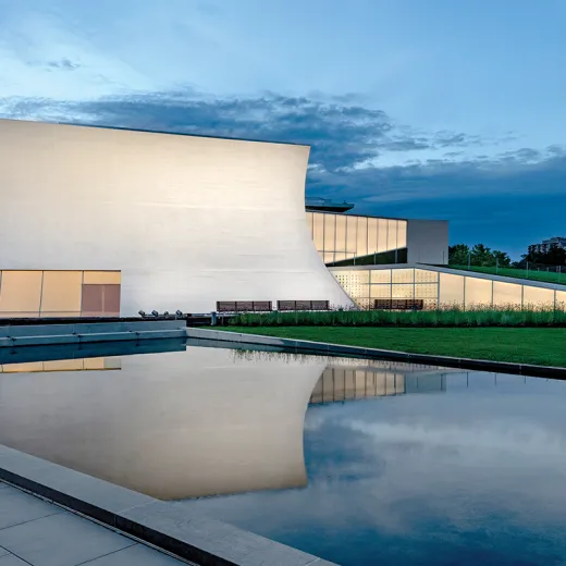 A white rectangular building, The Kennedy Center's Reach, is captured in a reflecting pool at dusk. 