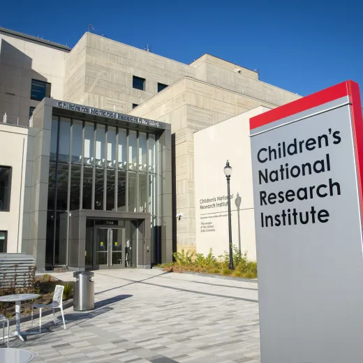 Signage for the Children's National Research Institute is displayed in the foreground, with the tan-colored building situated behind it under a blue sky.