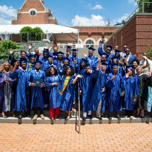 A group of smiling, waving graduates wearing blue caps and gowns stand on steps in front of a building at Georgetown University.