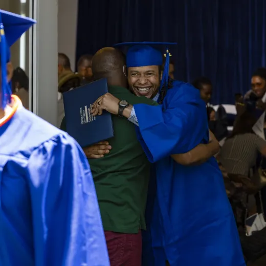 A smiling graduate in cap and gown hugs another person while proudly holding their diploma during a graduation ceremony.