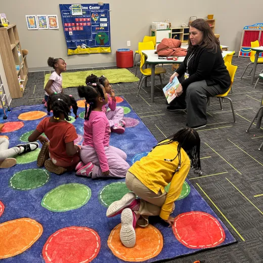 A teacher reads a book to students seated on a rug in a colorfully decorated classroom.