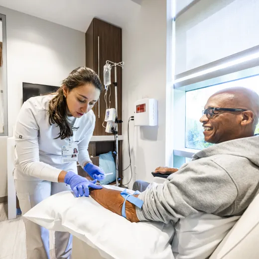 A healthcare professional prepares to draw blood from a patient's arm. The smiling patient is seated in a comfortable hospital room.