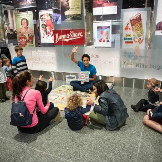 An group of elementary schoolchildren raise their hands to answer a question from a teacher. An engraver etches the names 'A. James and Alice B. Clark' into a stone wall at the National Gallery of Art. A collection of vintage World War II aircraft is suspended from the ceiling in the main gallery of the National WWII Museum. A museum educator leads an interactive learning session for enthusiastic parents and young children at a Smithsonian musem.