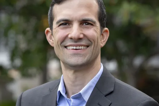 A professional headshot of Andrew Knutson. He wears a black blazer and a blue shirt.