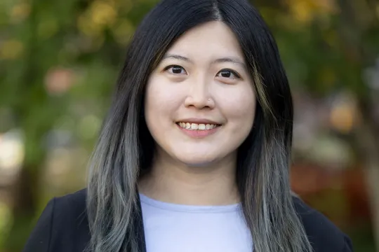 A professional headshot of Joan Zhang. She has long dark hair and wears a blue shirt and black sweater.