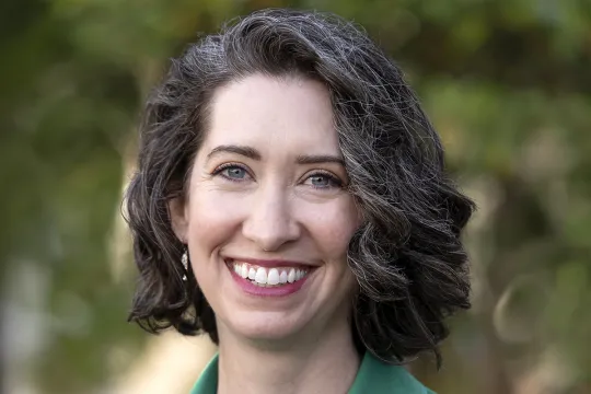 A professional headshot of Sarah Elbert. She has brown chin-length hair and wears a green dress.