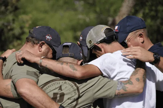 A group of men stand shoulder to shoulder and lock arms in a large group hug. Their faces are obscured.