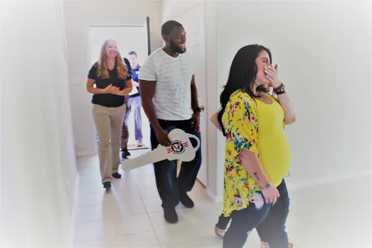 A mother-to-be gasps with joy as she enters her new home. Her husband follows her, holding a large key adorned with the Operation Homefront logo.