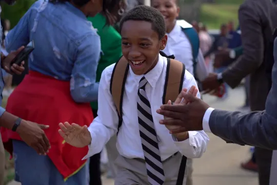 An elementary school student walks through a tunnel of adults who are cheering him on. The students and adults are wearing business attire.