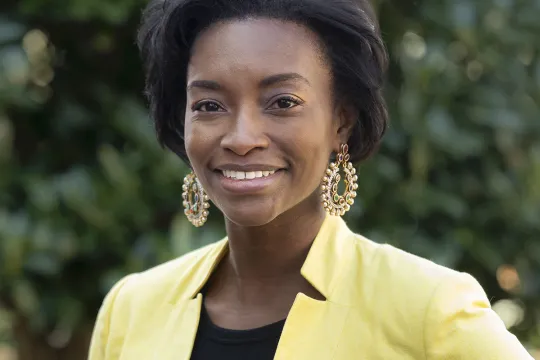 A professional headshot of Natalie Grandison. She has chin-length brown hair and wears a yellow blazer over a black shirt.