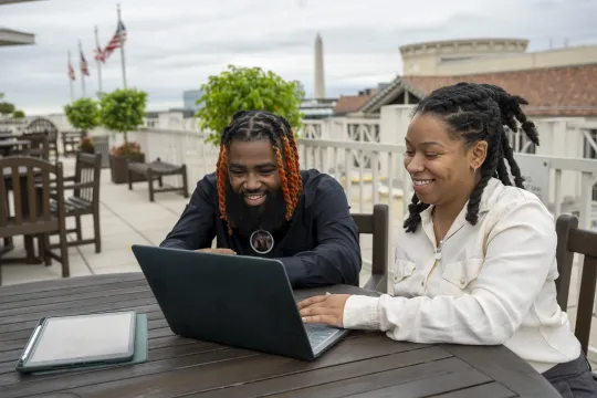 Two young people smile as they work on a laptop seated in an outdoor courtyard. The Washington Monument is visible in the skyline.