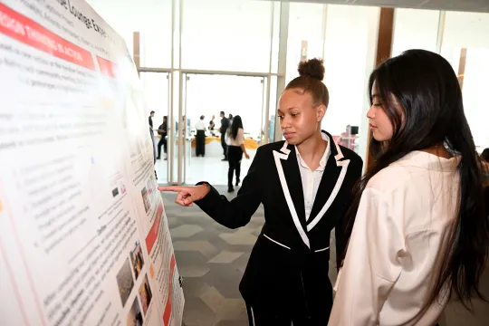 A student in business attire explains a presentation to another student while pointing at a poster board.