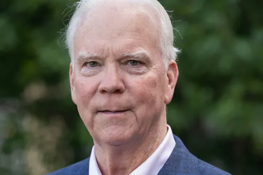A professional headshot of Board Director Robert Flanagan. He wears a blue blazer and a pink shirt.