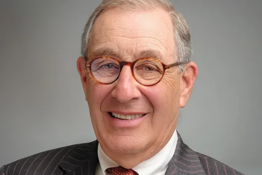 A professional headshot of Larry Nussdorf. He wears glasses, a grey blazer and an orange tie.