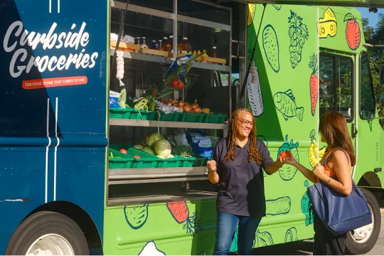 Two women stand outside a bright green grocery truck labeled "Curbside Groceries." One woman smiles at the other while she hands her a tomato.