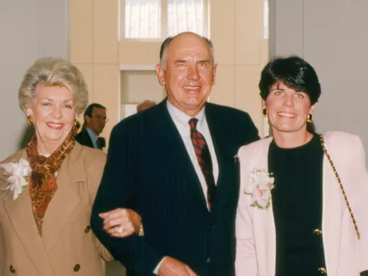 Mr. and Mrs. Clark stand arm in arm and smile at the camera with their daughter Courtney. They are all dressed in business attire.