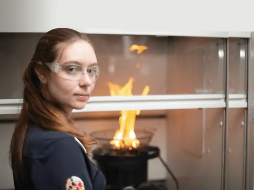 A doctoral student stands outside of a glass-enclosed fire. She wears protective goggles and looks confidently at the camera.