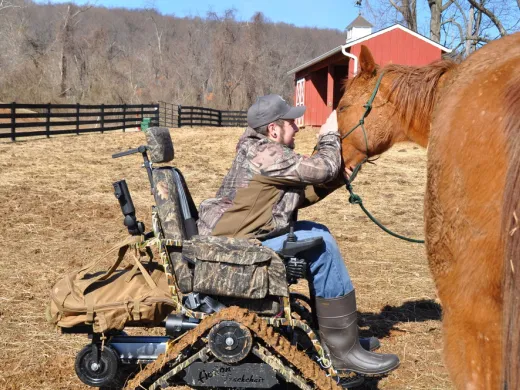 An army veteran in a specialized wheelchair pets the forehead of a chestnut horse.