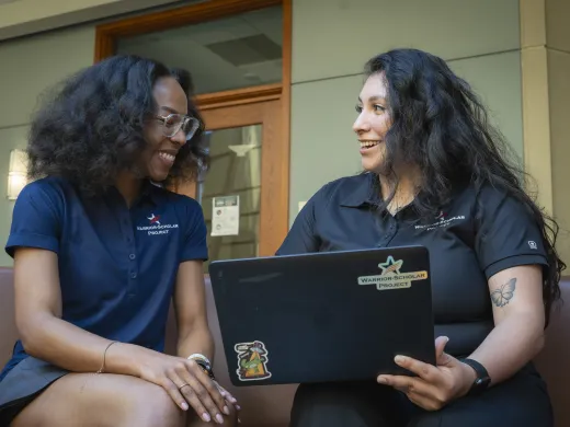 Two young women exchange smiles as they work on a laptop. Their blue polo shirts bear the Warrior-Scholar Project logo.