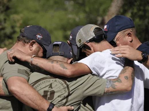 A group of men stand shoulder to shoulder and lock arms in a large group hug. Their faces are obscured.
