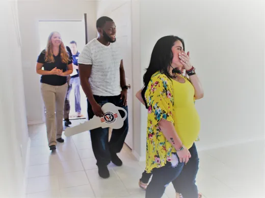 A mother-to-be gasps with joy as she enters her new home. Her husband follows her, holding a large key adorned with the Operation Homefront logo.