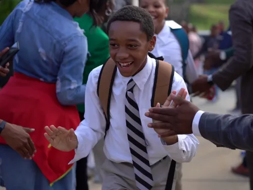An elementary school student walks through a tunnel of adults who are cheering him on. The students and adults are wearing business attire.