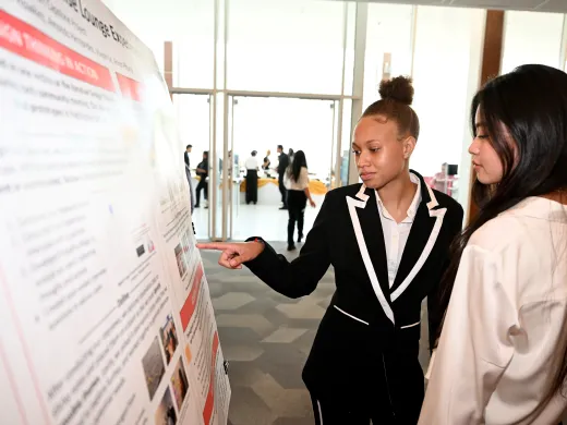 A student in business attire explains a presentation to another student while pointing at a poster board.