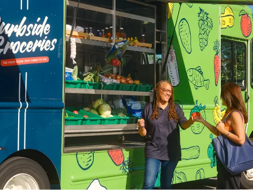 Two women stand outside a bright green grocery truck labeled "Curbside Groceries." One woman smiles at the other while she hands her a tomato.
