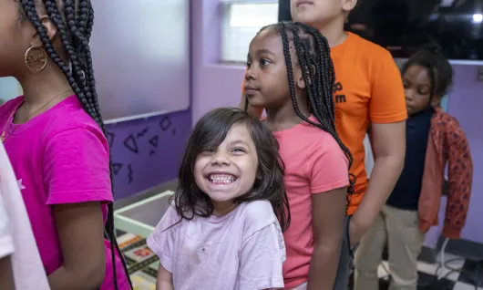 Children wait in line. A young girl, center, smiles eagerly at the camera.