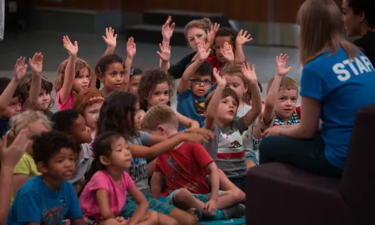 An group of elementary schoolchildren raise their hands to answer a question from a teacher.