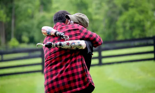 Two gentleman embrace in an outdoor, farm setting. One of the men has two prosthetic arms.