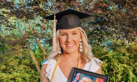 A young woman in a dress and a graduation cap holds a photograph of her father in his military uniform.