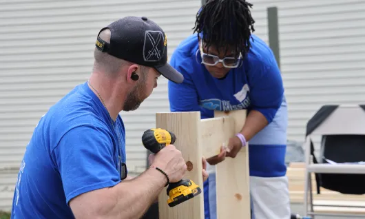 Volunteers from the Mission continues build materials together. A man, front drills, while a woman holds the lumber in place.