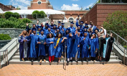 A group of smiling, waving graduates wearing blue caps and gowns stand on steps in front of a building at Georgetown University.
