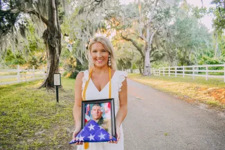 Katie Palmer smiles while holding a photo of her father in his military uniform. She is wearing her graduation regalia.