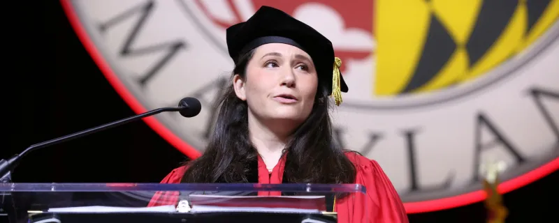 A woman in college regalia gives a speech at a podium. The University of Maryland seal is visible behind her on the stage.