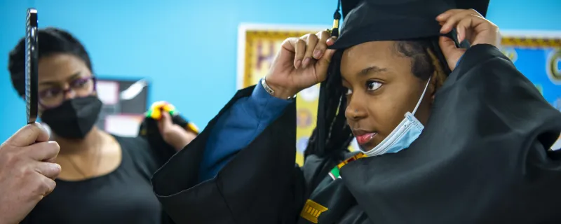 A high school student wearing graduation regalia tries on her cap while her mother looks on.