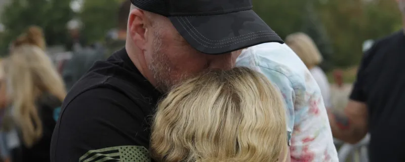 A man in a baseball cap with the Headstrong logo hugs a woman and kisses her on the top of her head.