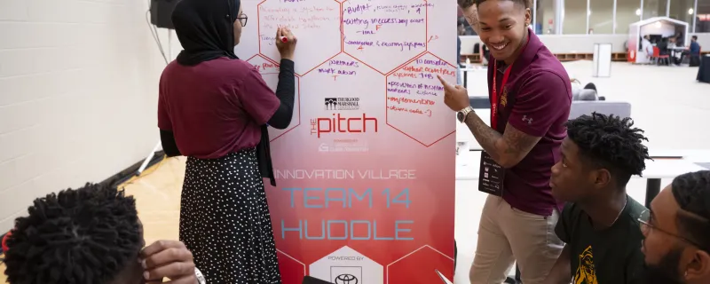 A group of five students gathered around a dry erase board jot down ideas.