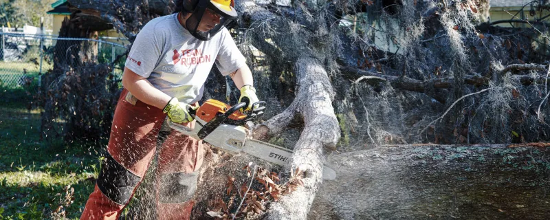 A Team Rubicon volunteer in a helmet and safety gear uses a chainsaw to cut up a large fallen tree.